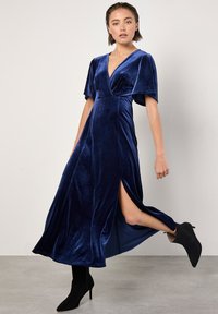 Robe maxi en velours bleu marine avec un décolleté en V profond, des manches flottantes et une fente sur le côté, associée à des bottines à talons noires.