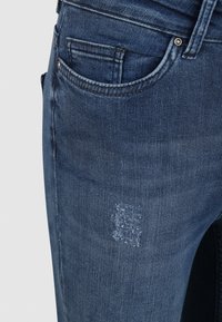 Vaqueros de denim azul oscuro con un diseño clásico de cinco bolsillos, un ligero desgastado y un parche desgastado en el muslo delantero.