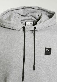 Sweat à capuche gris clair avec cordons noirs et un petit patch noir avec un design blanc minimaliste sur la poitrine.