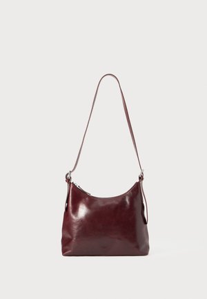 Burgundy leather shoulder bag with a curved shape, a zip closure, and a single adjustable strap. Smooth texture, minimalistic design.