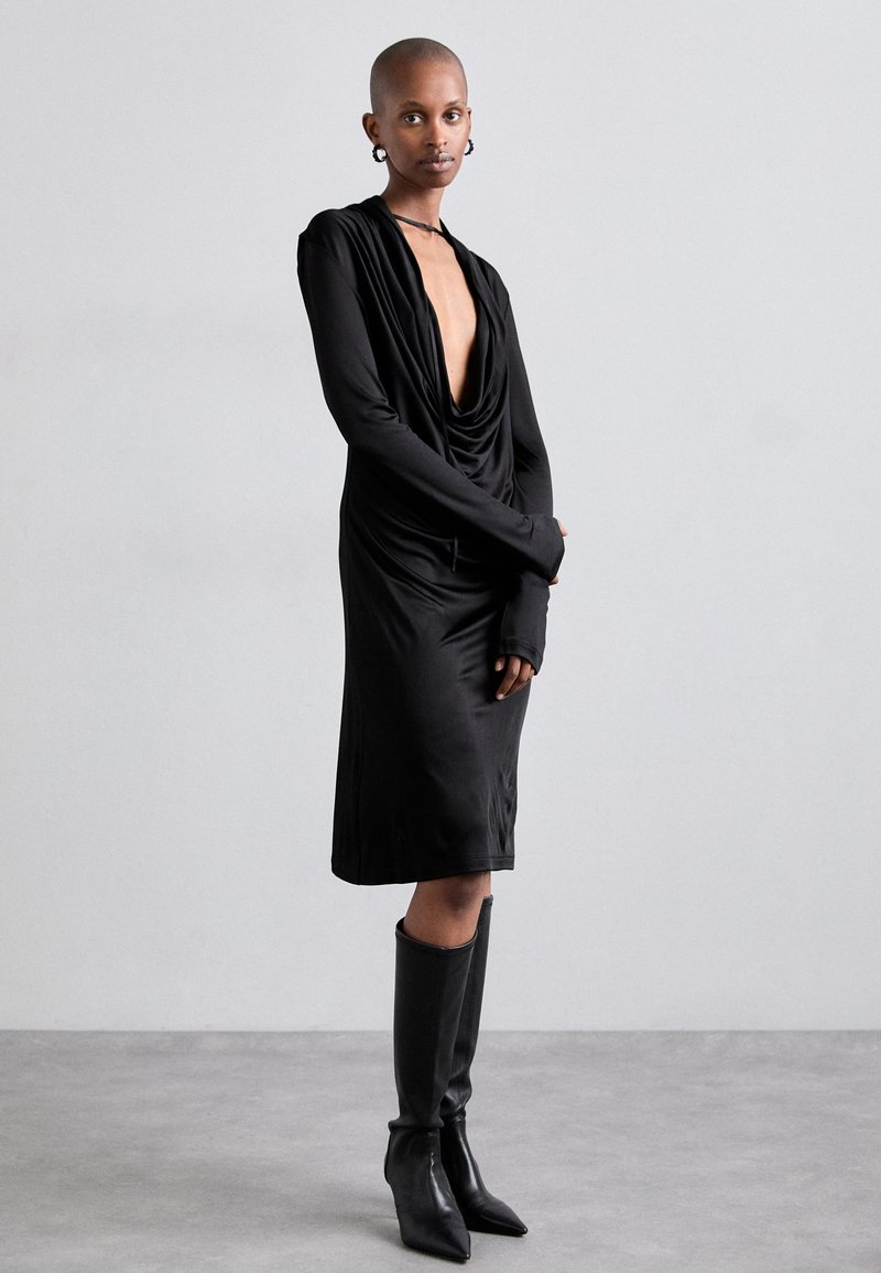 Black, draped long-sleeve dress with a deep V-neck, featuring fitted cuffs. Paired with knee-high pointed black boots. Smooth texture.