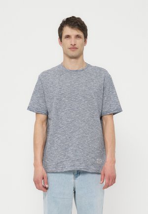 Άνδρας που φοράει ανοιχτό μπλε ριγέ κοντομάνικη μπλούζα και ανοιχτό μπλε τζιν, στέκεται μπροστά σε απλό λευκό φόντο.