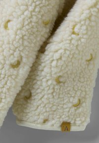 Krämfärgad fleecefilt med en texturerad yta som har små, broderade gula halvmånar och en beige etikett.