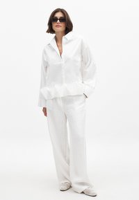 White button-up shirt with a ruffled hem, paired with loose white pants and textured loafers. The outfit is complemented by dark sunglasses.