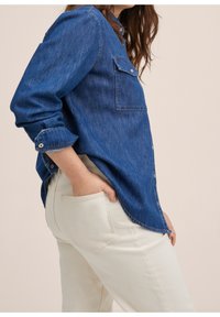 Camisa de mezclilla de manga larga, color azul, con botones en la parte delantera, un bolsillo en el pecho, llevada con pantalones color crema, la persona tiene una mano en el bolsillo.