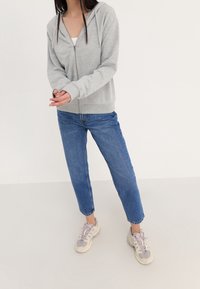 Sudadera gris con cremallera y capucha, combinada con jeans rectos de color azul claro y zapatillas ligeras. Tejido suave, corte relajado y diseño sencillo.