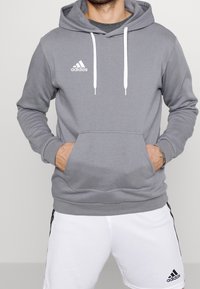 Homme portant un sweat à capuche Adidas gris avec des cordons blancs et un short Adidas blanc avec des bandes noires sur les côtés. Mains dans les poches du sweat à capuche.