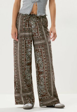 Kobieta nosząca luźne brązowe spodnie z szerokimi nogawkami i wzorem, z sznurkiem w talii, w połączeniu z krótkim szarym topem bez rękawów i casualowymi butami.