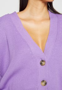 Paarse gebreide cardigan met een diepe V-hals, geribbelde textuur en twee grote, gegoten knopen aan de voorkant.