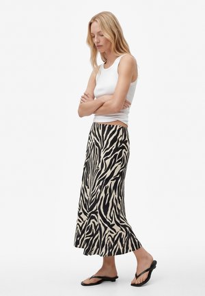 Femme aux cheveux longs et blonds, portant un haut blanc sans manches, une jupe midi à imprimé zèbre noir et beige, et des tongs noires, debout les bras croisés.