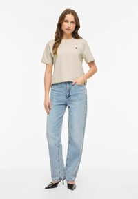 Beżowy bawełniany t-shirt z krótkimi rękawami i czarnym logo serca na piersi, zestawiony z jasnoniebieskimi, wysokim stanem dżinsami oraz czarnymi szpilkami.