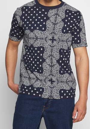Mand iført navyblå t-shirt med hvidt paisley- og prikmønster og mørkeblå jeans, stående med den ene hånd i lommen.