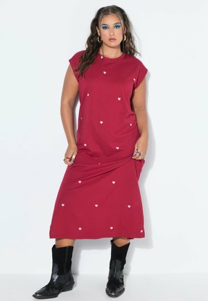 Femme aux longs cheveux foncés portant une robe rouge avec de petits cœurs blancs et des bottes de cow-boy noires, debout devant un fond blanc.