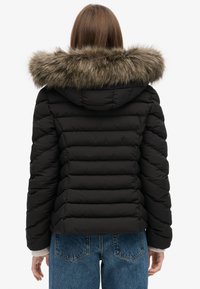 Zwarte pufferjack met een horizontaal gewatteerd ontwerp, voorzien van een capuchon met nepbontvoering en geribde manchetten, gedragen over hoog-getailleerde jeans.