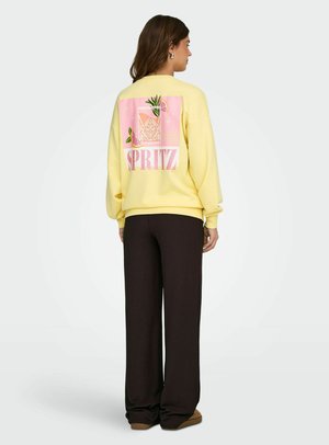 Vrouw draagt een gele trui met een roze drankje en de tekst "SPRITZ" op de achterkant, gecombineerd met losse zwarte broek en bruine schoenen, staand opzij.