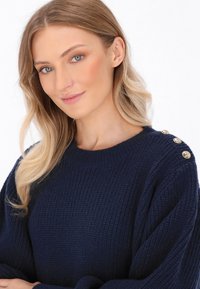 Marine gebreide trui met geribbelde textuur, oversized mouwen en gouden knoopaccenten op de schouder. Zachte stof en ronde halslijn.