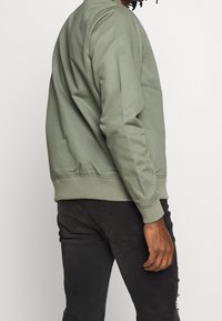 Zielona kurtka bomberka z prążkowanym kołnierzem i mankietami, cechująca się gładką, matową fakturą. Połączona z ciemnymi dżinsami, podkreślającymi swobodny wygląd.