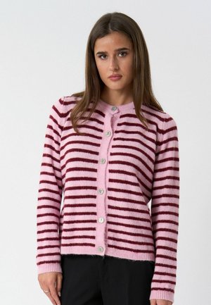 Kobieta o prostych brązowych włosach, ubrana w różowo-czerwony, poziomo paskowany kardigan na guziki oraz czarne spodnie, stojąca na tle jednolitego tła.