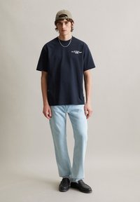 Marineblauw katoenen t-shirt met korte mouwen, voorzien van witte tekst op de borst. Gecombineerd met lichtblauwe rechte jeans en zwarte schoenen.