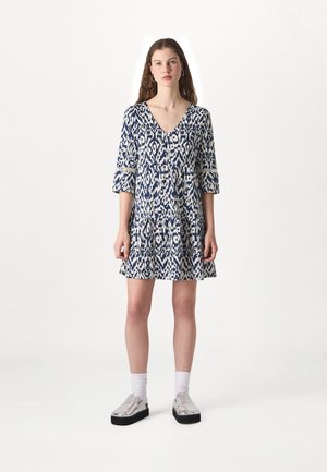 Abito blu e crema con motivo, scollatura a V profonda, maniche a tre quarti e gonna a ruota. La modella indossa scarpe slip-on argentate.
