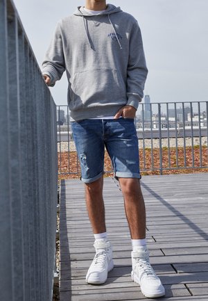 Šedá mikina s kapucí a předním kapsou, modrý grafický detail; oprané modré džínové šortky s ohrnutými lemy; bílé vysoké tenisky, stojící na dřevěné terase.