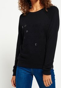 Donna che indossa un felpa nera con scritte in rilievo che elencano città: Parigi, New York, Londra, abbinata a jeans blu.