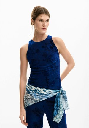 Tmavě modrý top bez rukávů s květinovou texturou, doplněný světle modrým a bílým šátkem s vzorem uvázaným v pase. Hladká látka, přiléhavý střih.