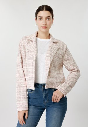 Tumejuustega naine kannab heleroosa ja valge tekstuuriga jopet valge särgi ja siniste teksade peal, seistes ühevärvilise tausta ees.