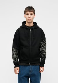 Jauns vīrietis valkā melnu kapuci ar rāvējslēdzēju, uz kuras abām piedurknēm ir baltas zirnekļa tīkla dizaini, un zilu džinsu bikses, stāvot pret vienkāršu fona.