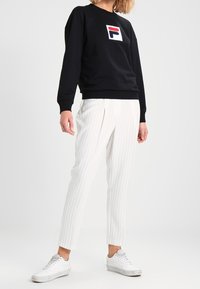 Persona vestida con sudadera negra con logotipo, pantalones a rayas blancas y zapatillas blancas, de pie frente a un fondo liso.