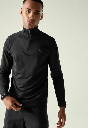 Young man wearing a black long-sleeve athletic top with a quarter zip and a smartwatch on his left wrist against a plain background.