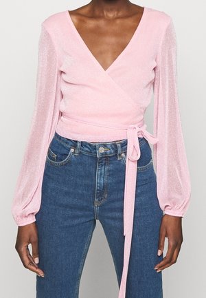 Woman wearing a shimmery pink wrap top with long puffed sleeves and denim high-waisted jeans, front view cropped at shoulders.