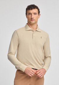 Beżowa koszulka polo z długim rękawem wykonana z miękkiego materiału, z kołnierzykiem, zapięciem na guziki i małym logo na lewej piersi. Gładka struktura.