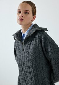 Zesílený svetr s copánkovým pletením v tmavě šedé barvě, s vysokým límcem, polovičním zipem a žebrovanými prvky na výstřihu a rukávech.