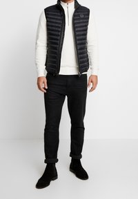 Gilet imbottito nero con texture trapuntata sopra una maglietta a maniche lunghe color crema, abbinato a jeans neri e stivaletti alla caviglia in suede neri.