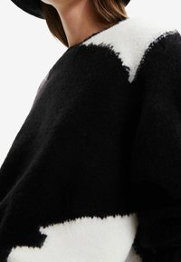 Czarnobiały dzianinowy sweter o teksturowanym wykończeniu, z wyrazistym wzorem gwiazdy i luźnym krojem, częściowo przykryty czarnym kapeluszem.