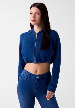 Frau steht in einer blauen, kurzen Jeansjacke mit Reißverschluss und hoch taillierten blauen Jeans und blickt leicht nach links vor einem schlichten, hellen Hintergrund.