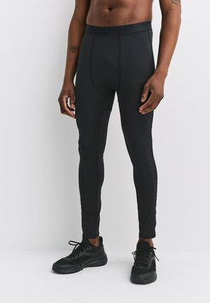 Man wearing black athletic leggings and black running shoes, standing against a plain white background, shirtless.