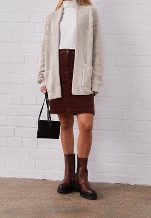 Osoba nosící béžový pletený svetr, bílý rolák, hnědou sukni, hnědé kotníkové boty, držící malou černou kabelku na pozadí bílé cihlové stěny.