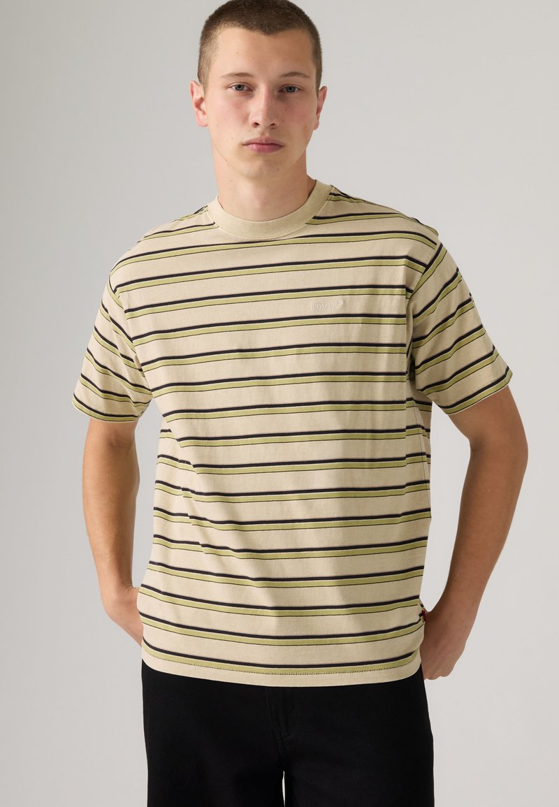 Jeune homme portant un t-shirt beige à manches courtes avec des rayures horizontales noires et olive, debout devant un fond clair uni.
