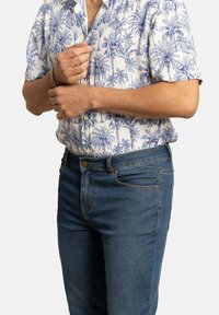 Camisa de manga corta en blanco con estampados de palmeras azules; combinada con jeans azul oscuro con cierre de botón y bolsillos frontales.
