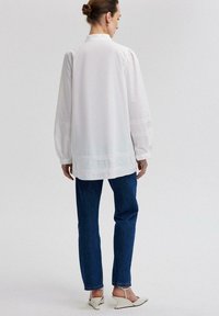 Blusa bianca a maniche lunghe con colletto alto, presenta maniche arricciate e cuciture decorative all'orlo, indossata con jeans scuri e scarpe chiare.