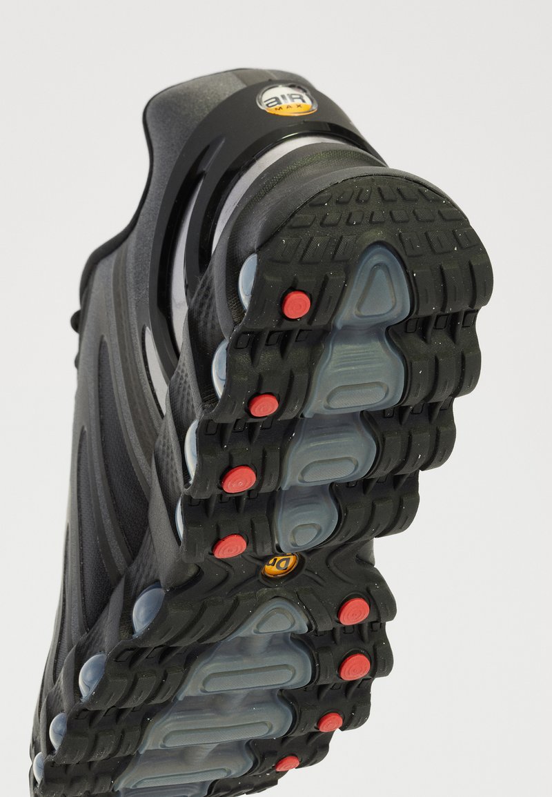 Zapatilla deportiva negra con una suela de goma texturizada, amortiguación de aire visible, detalles en rojo y un diseño de talón redondeado.