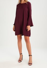 Kastanjebrun A-linje klänning med långa klockärmar, rund hals och mjukt tyg. Buren med svarta sandaler med remmar, mot en enhetlig bakgrund.