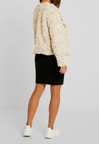 Valkoinen tekoturkistakki, jossa on aaltoileva kuvio ja lyhyt malli, yhdistetty mustaan kynähameeseen ja valkoisiin tennareihin, kuvattu takaa päin.