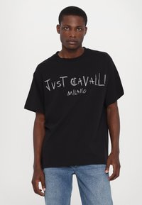 Melnas kokvilnas T-krekls ar baltiem izdrukātiem burtiem "JUST CAVALLI MILANO" priekšpusē. Brīvs piegriezums ar īsām piedurknēm. Piemērots ikdienas valkāšanai.