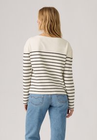 Gestreifter Pullover in Creme und Schwarz mit langen Ärmeln und lässiger Passform, kombiniert mit hochgeschnittenen blauen Jeans, von hinten betrachtet.