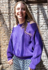 Jongere vrouw die in het zonlicht lacht tegen een bakstenen muur, gekleed in een paarse gebreide trui met uitsnijdingen op de schouders en bedrukt een broek.