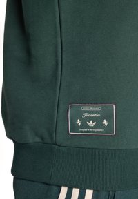 Mørkegrøn sweatshirt med ribbet tekstur, lodrette sømdetaljer og et broderet mærke med brandinformation.