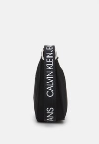 Czarna nylonowa torba z grubym, teksturowanym paskiem z białym logo. Design jest prosty, o prostokątnym kształcie i minimalnej ilości okuć.